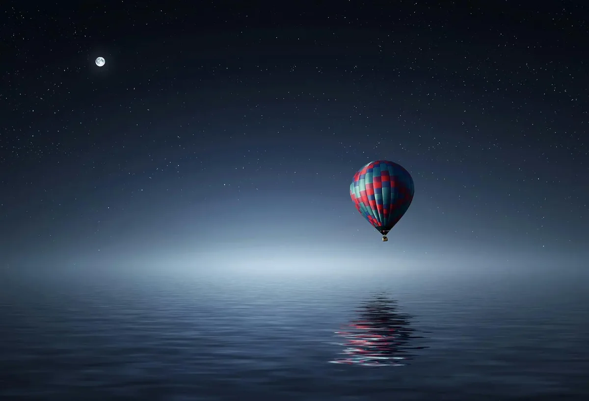 Single hot air balloon floating over calm water under a moonlit starry sky