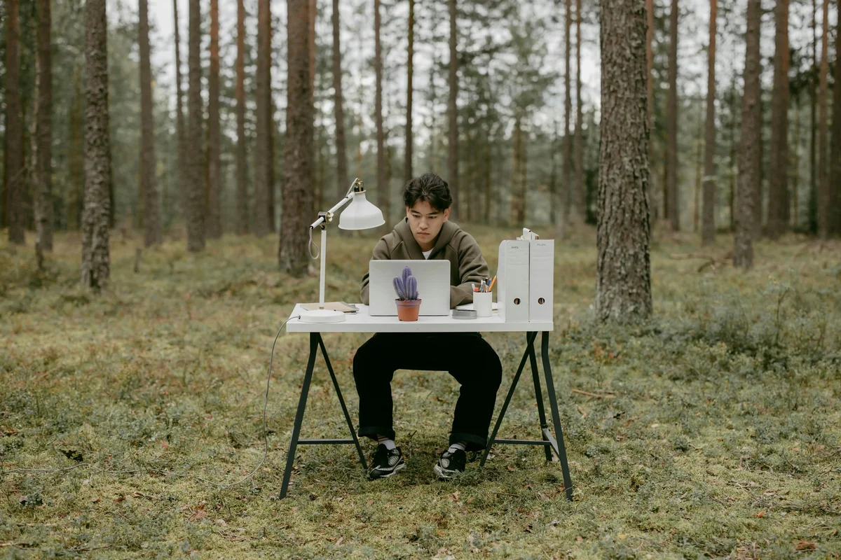 Person working at a white desk with a laptop and lamp in a forest clearing