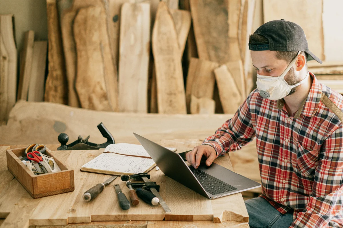 Man in a cap and dust mask using a laptop at a wood workshop bench