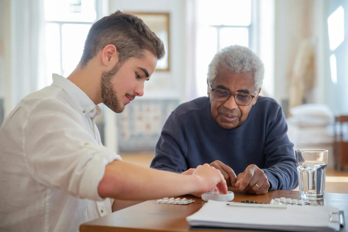 Young man helping an older man sort pills into a medication organizer at a table