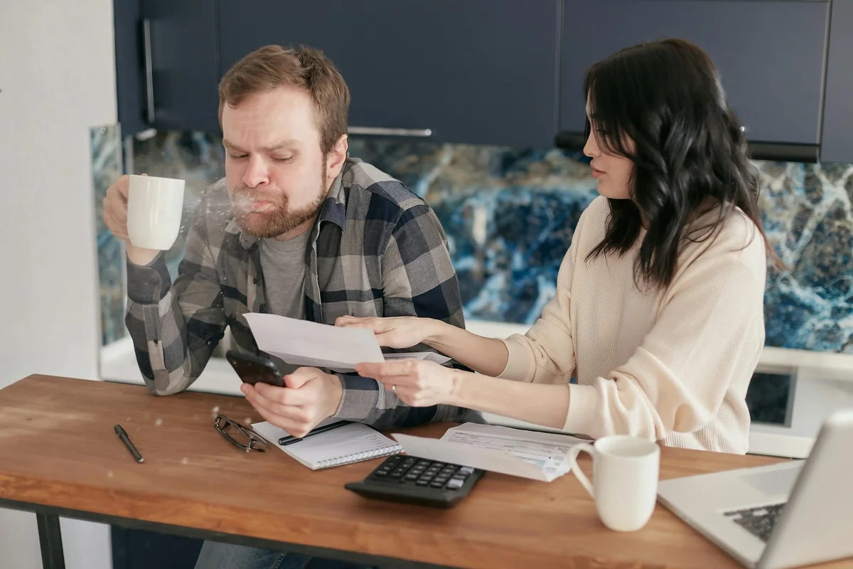 Man spitting out coffee in shock as a woman shows him a bill at a kitchen table