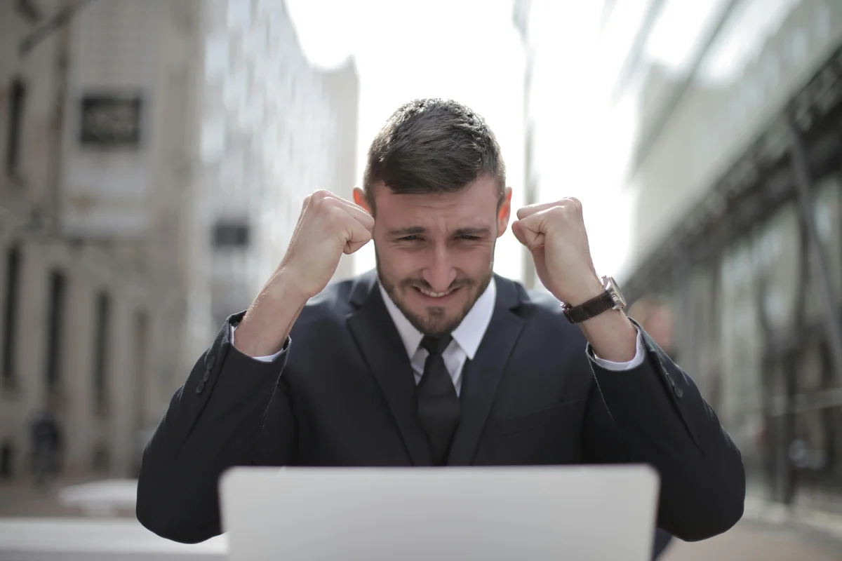 Man in a dark suit clenching his fists at a laptop on a city street