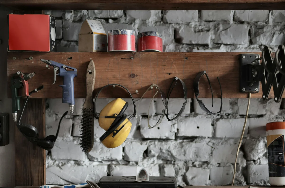 Safety glasses, ear protection, and tools hanging on a workshop rail above a brick wall