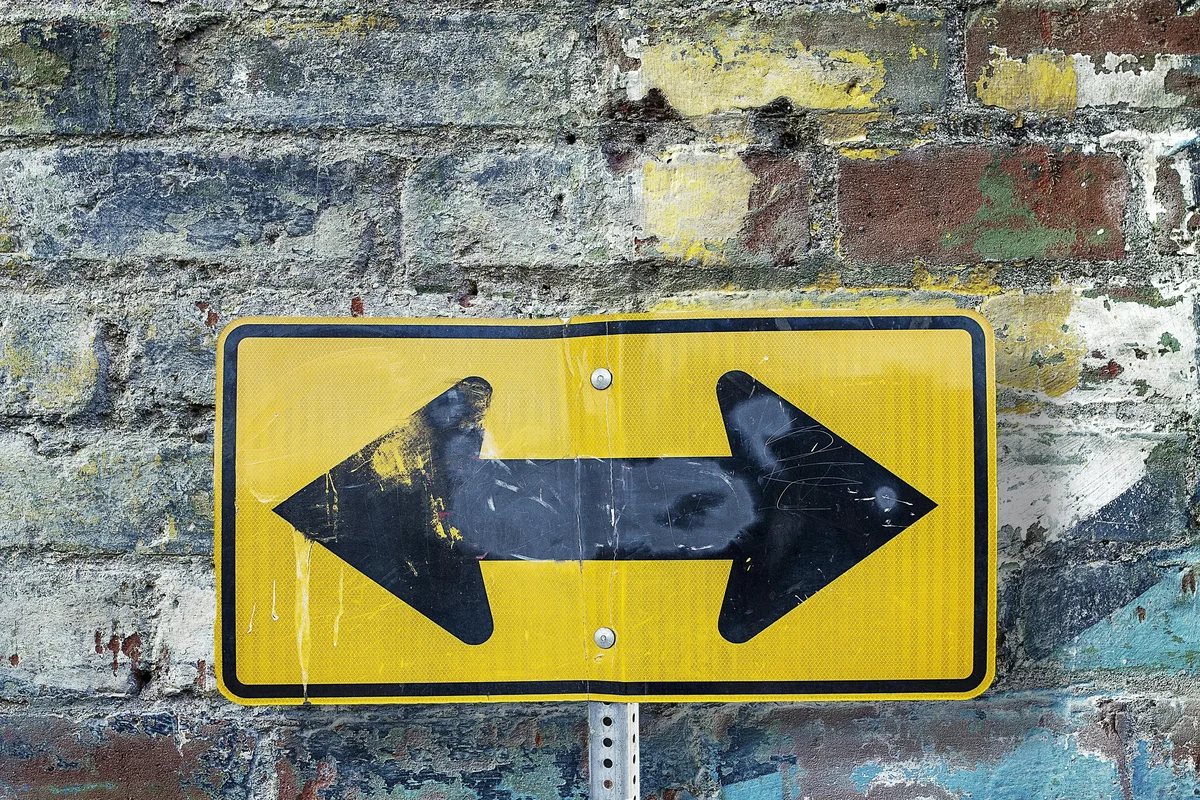 Worn yellow double-headed arrow sign on a graffiti-covered brick wall