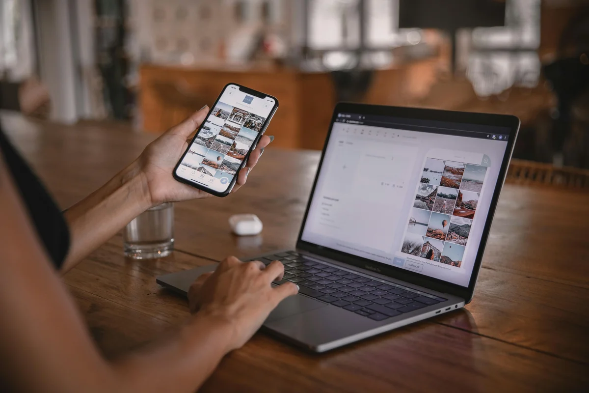 Person holding a phone with a photo gallery beside an open laptop on a wooden desk