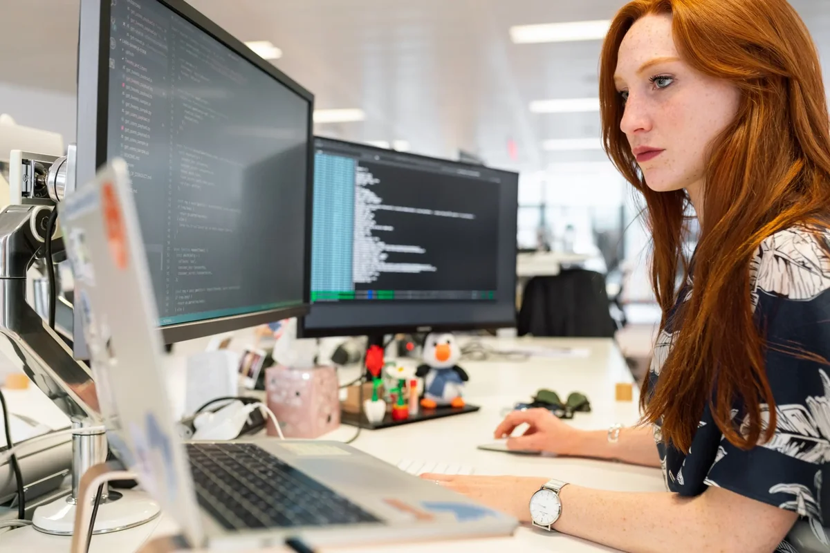 Red-haired woman writing code at a desk with dual monitors